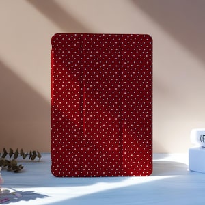 May include: A red tablet case with a white polka dot pattern. The case is rectangular and stands upright. The background includes flowers and books.
