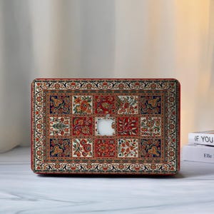 Puede incluir: Una funda para portátil con un diseño vibrante que se asemeja a una alfombra persa. La funda presenta un borde rojo y un diseño central con motivos florales y geométricos en rojo, azul y dorado. El logotipo de Apple es visible.