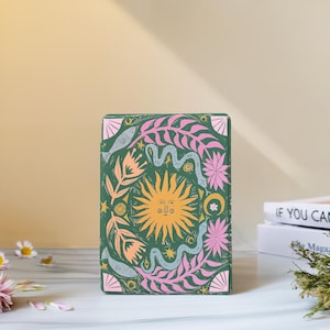 May include: A rectangular green book cover featuring a central yellow sun with a face, surrounded by pink leaves, blue snakes, fish, and flowers. The design includes small stars and a crescent moon, creating a detailed, colorful illustration on the front surface.