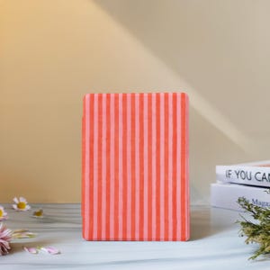 May include: A rectangular, coral-colored case with vertical stripes in a lighter shade of coral. The case is standing upright on a light-colored surface. The background is a solid, light beige color.