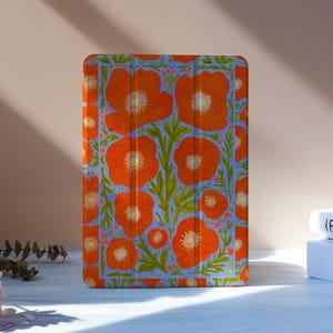 Puede incluir: Una funda para tableta con un estampado floral en naranja y azul. El diseño presenta flores naranjas y hojas verdes sobre un fondo azul claro. La funda es rectangular y tiene una cubierta plegable.