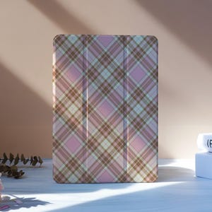 May include: A pink, brown, and white plaid tablet case stands upright. The case is rectangular with a folding cover. The background includes flowers and books. The case is designed to protect a tablet.