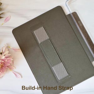 May include: A dark olive green tablet case featuring a rectangular centre cutout with a grey elastic hand strap secured by stitching. The case includes a clear plastic edge component and the text Build-in Hand Strap printed at the bottom.