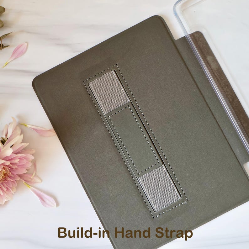 May include: A dark olive green tablet case featuring a rectangular centre cutout with a grey elastic hand strap secured by stitching. The case includes a clear plastic edge component and the text Build-in Hand Strap printed at the bottom.