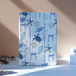 May include: A light blue tablet case featuring a repeating pattern of marine life. The design includes blue starfish, seashells, turtles, and bows. The case is standing upright on a white surface, with a soft pink background.