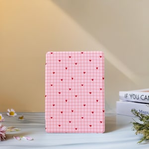 Puede incluir: Un cuaderno rectangular con un patrón de cuadrícula rosa y pequeños corazones rojos dispersos. El cuaderno está de pie sobre una superficie de color claro. El fondo es beige neutro.