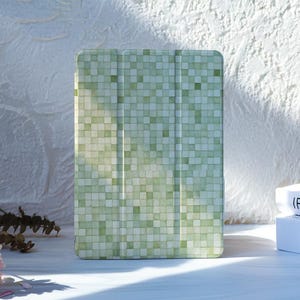 Puede incluir: Una funda rectangular para tableta con un patrón de mosaico en tonos verdes y blancos. La funda tiene un diseño de tres pliegues con líneas verticales y un acabado liso. El patrón consta de pequeños cuadrados variados en tonos salvia, oliva y blanco.