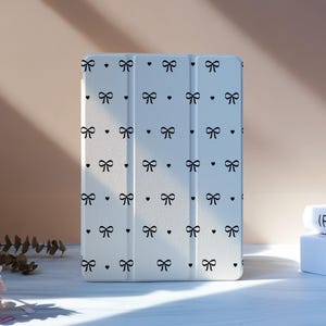 以下が含まれることがあります： 黒いリボンとハートの繰り返し模様が施された白いタブレットケース。ケースは白い面に立っています。背景には2冊の本とピンクの花があります。