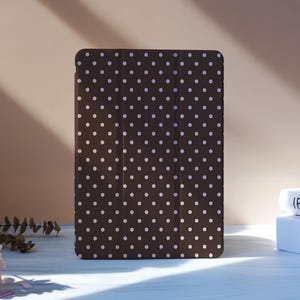 Puede incluir: Una funda marrón para tableta con un diseño de lunares blancos. La funda está de pie sobre una superficie blanca, con flores rosas y libros en el fondo. La funda está diseñada para proteger una tableta.