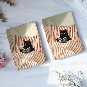 May include: Two rectangular e-reader covers featuring an illustration of a black cat lying in bed under red and white striped blankets. The cat is holding an open book with a floral pattern on the cover. The background is a soft cream colour.