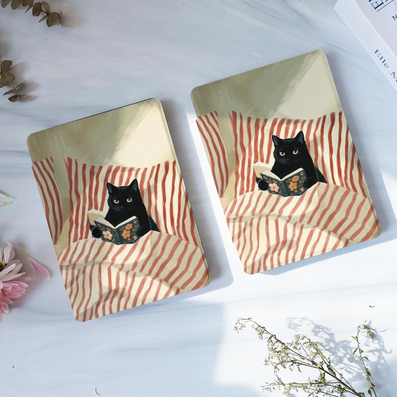 May include: Two rectangular e-reader covers featuring an illustration of a black cat lying in bed under red and white striped blankets. The cat is holding an open book with a floral pattern on the cover. The background is a soft cream colour.