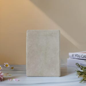 Puede incluir: Un diario o cuaderno rectangular gris claro con una cubierta texturizada se encuentra en posición vertical. El libro está sobre una superficie blanca con flores dispersas y una pila de libros en el fondo.