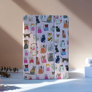 Puede incluir: Una funda para tableta de color rosa claro con un patrón repetido de gatos de dibujos animados en varias poses y atuendos. Los gatos son de diferentes colores, incluyendo negro, gris, naranja y blanco. La funda está de pie.