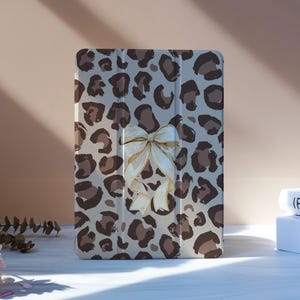 Puede incluir: Una funda para tableta con estampado de leopardo en tonos marrón y beige. Un lazo crema con ribete dorado está centrado en la parte delantera. La funda está de pie sobre una superficie blanca.