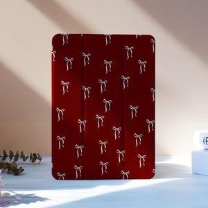 Puede incluir: Una funda de tableta burdeos con un patrón repetido de lazos blancos. La funda está de pie sobre una superficie blanca, con libros y flores rosas en el fondo. La funda está diseñada para proteger una tableta.