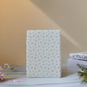 Puede incluir: Una funda rectangular blanquecina con un delicado estampado de pequeñas flores azules. La funda está de pie sobre una superficie clara. Parece ser una funda protectora para un dispositivo electrónico.