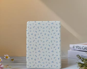 May include: An off-white rectangular case, covered in a pattern of tiny blue floral motifs. The case is standing on a light surface. It appears to be a protective cover for an electronic device, such as a tablet or e-reader.