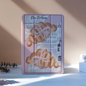 Pode incluir: Uma capa rosa para tablet com uma ilustração em aquarela de croissants amarrados com fitas rosa. O fundo é uma impressão de jornal com o texto "The Tribun" e "Corner Cafe". A capa está sobre uma superfície branca.