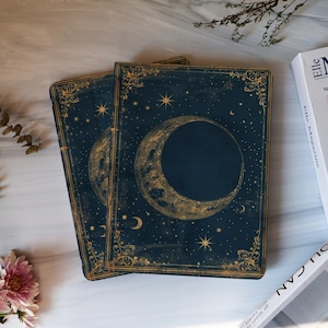 May include: Two dark blue journals with gold crescent moon and star designs. The covers have ornate gold borders and a celestial theme. The journals are stacked on a marble surface, with a magazine and flowers in the background.