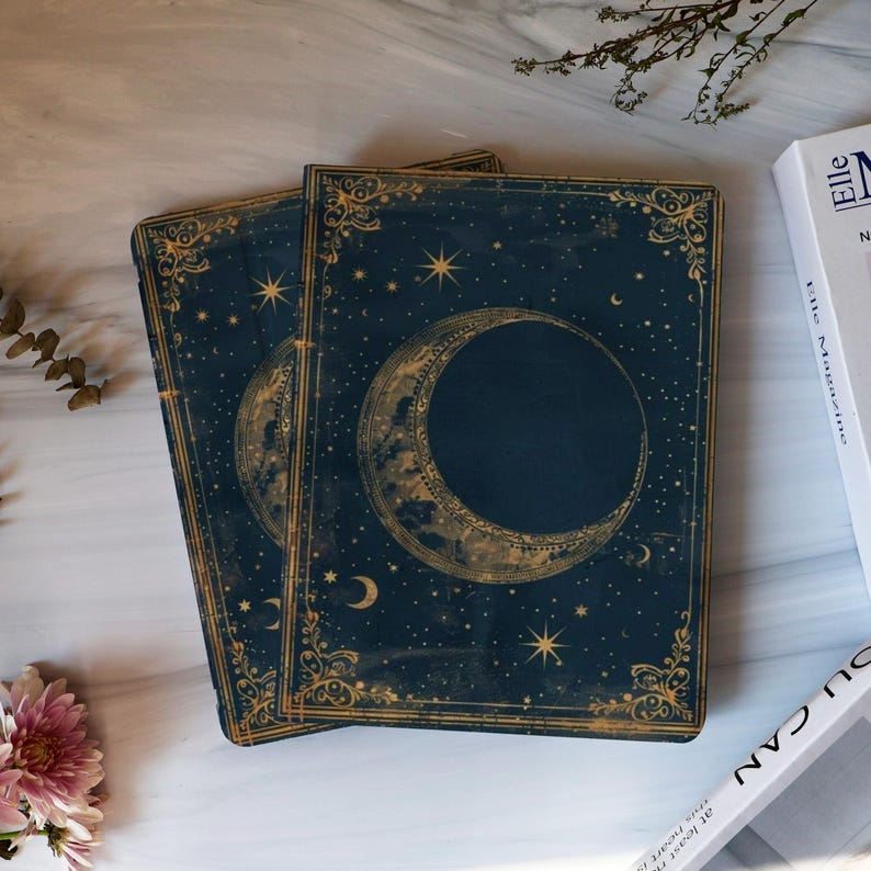 May include: Two dark blue journals with gold crescent moon and star designs. The covers have ornate gold borders and a celestial theme. The journals are stacked on a marble surface, with a magazine and flowers in the background.