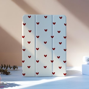 Puede incluir: Una funda blanca para tableta con un patrón de pequeños corazones rojos. La funda está de pie sobre una superficie blanca, con un arreglo floral rosa y dos libros en el fondo.