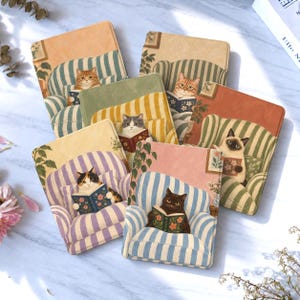 Puede incluir: Una colección de seis fundas rectangulares para lectores electrónicos, cada una con la ilustración de un gato sentado en un sillón a rayas leyendo un libro. Los gatos aparecen en varios colores y patrones, sobre fondos de diferentes tonos pastel.