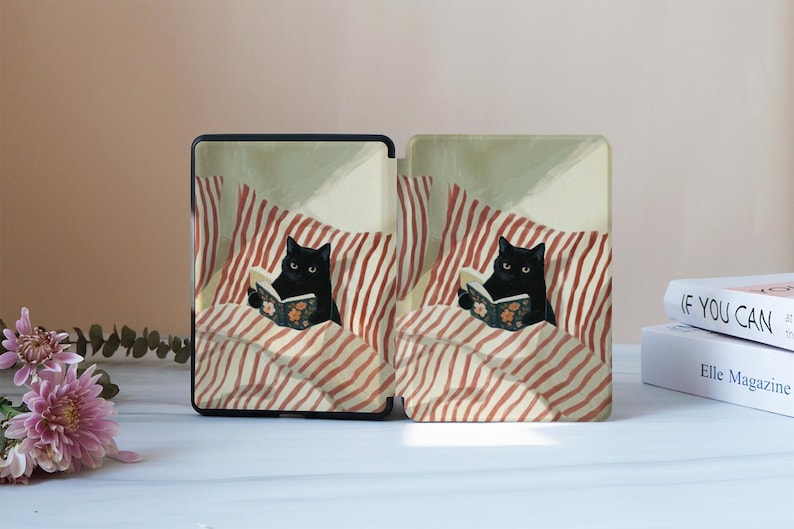 May include: A protective e-reader case featuring a printed illustration of a black cat reading a book whilst tucked into bed with red and white striped bedding. The design shows the cat holding an open book with a floral pattern on the cover.