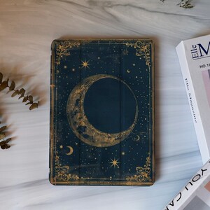 May include: Dark blue tablet case featuring a celestial design with a crescent moon and stars. The case has a vintage aesthetic with gold-colored decorative borders. The design is reminiscent of a night sky.