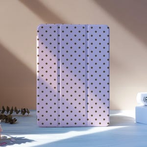 May include: A lavender tablet case with a repeating pattern of dark brown polka dots. The case is standing upright on a white surface, with a stack of books and flowers in the background.
