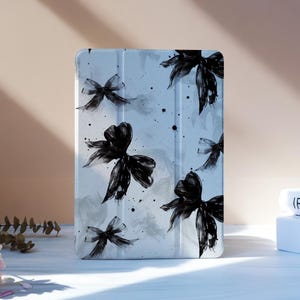 Puede incluir: Una funda para tableta azul claro con un diseño de acuarela de lazos negros y salpicaduras. La funda está de pie sobre una superficie blanca, con flores rosas y libros en el fondo.