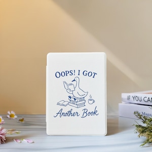 Może przedstawiać: Białe etui w kształcie książki z niebieskim napisem "OOPS! I GOT Another Book". Projekt przedstawia kaczkę czytającą książkę na stosie książek, z filiżanką kawy. Etui jest prawdopodobnie przeznaczone do czytnika e-booków lub tabletu.