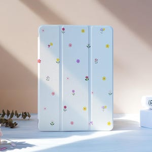 Peut inclure: Étui de tablette blanc orné d'un motif floral délicat rose, jaune et violet. L'étui est fermé, présentant un design épuré et minimaliste. Le fond présente une palette de couleurs douces et neutres, avec des livres et des fleurs.