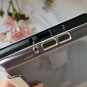 May include: A protective tablet case featuring a black inner frame and a clear, transparent outer shell. The top edge displays precise cutouts for the power button, speakers, and charging port, ensuring full access to all device functions.