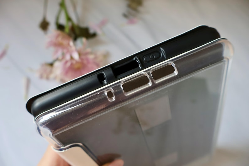 May include: A protective tablet case featuring a black inner frame and a clear, transparent outer shell. The top edge displays precise cutouts for the power button, speakers, and charging port, ensuring full access to all device functions.