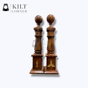 May include: Two dark brown wooden decorative pillars with a ball-shaped top and a square base. Each pillar has a decorative brass emblem on the front. The pillars are from The Kilt Corner.