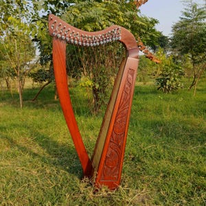 May include: A wooden harp with a reddish-brown finish stands in a grassy area. The harp features strings, tuning pegs, and decorative carvings on the soundbox. The instrument is designed with a curved neck and a triangular frame.