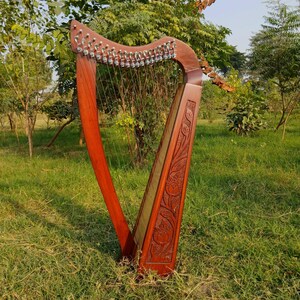 May include: A wooden harp with a rich, reddish-brown finish. The instrument features numerous strings and intricate carvings on the soundbox. The harp is set outdoors in a grassy area, showcasing its elegant design.