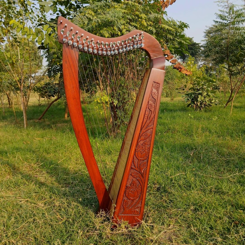 May include: A wooden harp with a rich, reddish-brown finish. The instrument features numerous strings and intricate carvings on the soundbox. The harp is set outdoors in a grassy area, showcasing its elegant design.