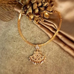 May include: A gold-toned necklace with a pendant featuring a fan-shaped design adorned with clear gemstones. The necklace has a delicate chain and is displayed on a textured surface with a golden floral accent.