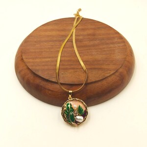 May include: A gold-toned necklace with a round pendant featuring green leaves, a pearl, and other embellishments. The pendant is set in a gold frame and hangs from a delicate chain. The necklace is displayed on a wooden surface.