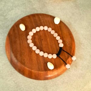 May include: A rose quartz bead bracelet with a black adjustable cord, on a wooden bowl. The bracelet has small, round, light pink beads. Seashells are scattered around the bowl.