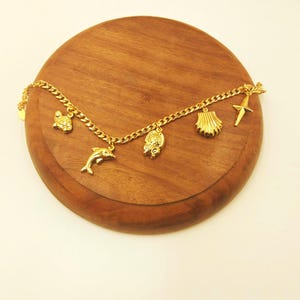 Può includere: Un braccialetto con ciondoli dorato con vari ciondoli a tema marino, tra cui un delfino, una tartaruga, una conchiglia e una stella marina. Il braccialetto è esposto su una superficie rotonda di legno, mostrando il suo design intricato e il tema nautico.