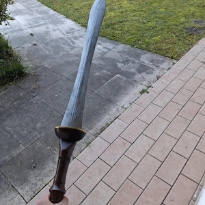 A person holding a large gray double-edged sword outdoors above a tiled patio and grass lawn.