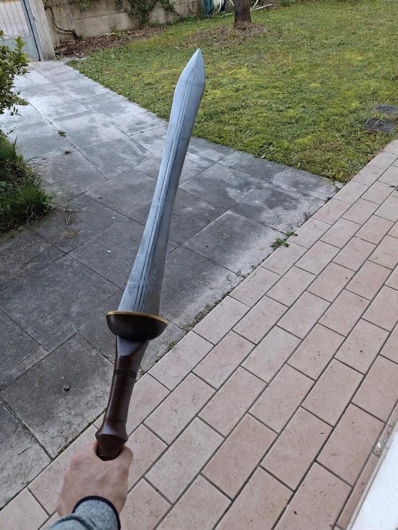 A person holding a large gray double-edged sword outdoors above a tiled patio and grass lawn.
