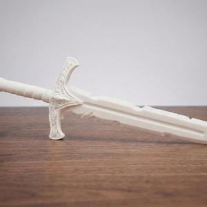 A plain white unpainted 3D printed short sword replica resting horizontally on a wooden table against a light background.