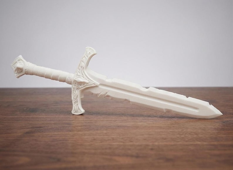 A plain white unpainted 3D printed short sword replica resting horizontally on a wooden table against a light background.