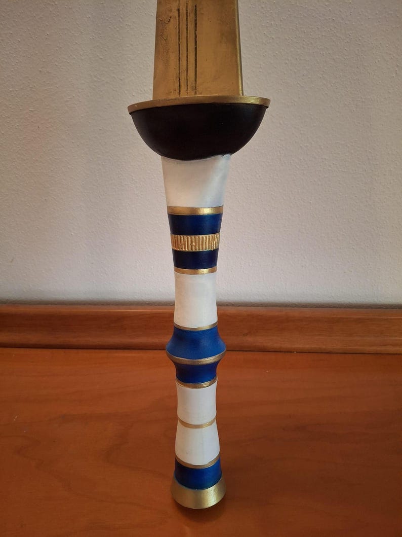 Close-up view of the decorative hilt of a fantasy sword with alternating white, blue, and gold rings and a dark bowl-shaped guard below the golden blade.