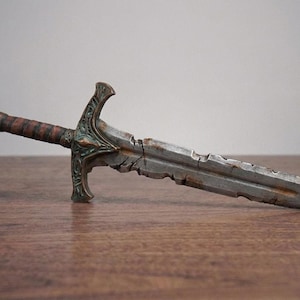 A weathered, battle-damaged short sword replica with metallic silver blade and brown hilt resting horizontally on a wooden surface.
