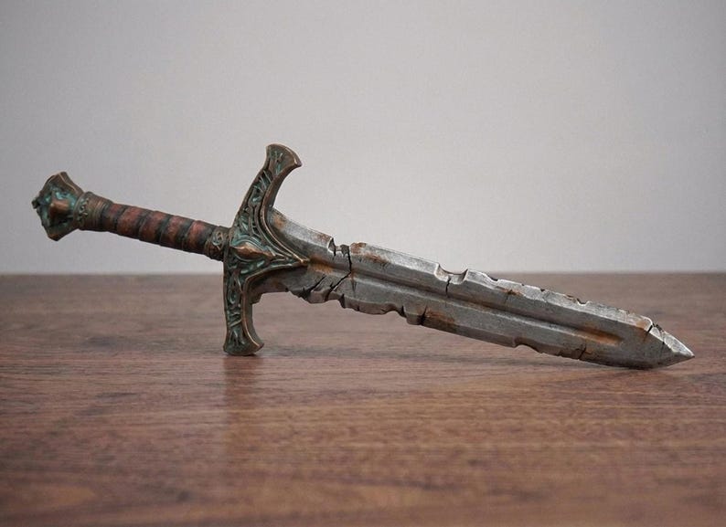 A weathered, battle-damaged short sword replica with metallic silver blade and brown hilt resting horizontally on a wooden surface.