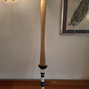 A tall fantasy sword with a golden blade and a hilt featuring white, blue, and gold bands standing vertically against a white wall.